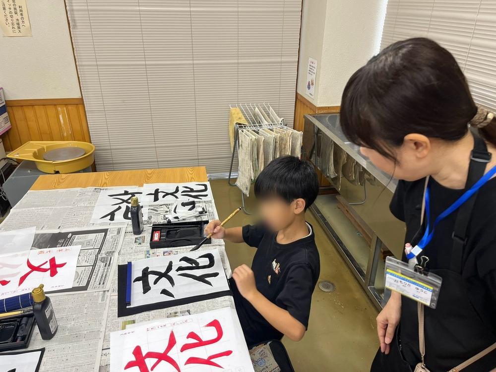 先生が朱墨で書いた字のよこで、筆で練習する男の子